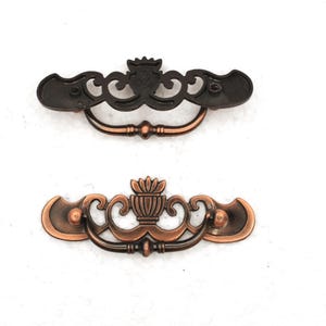 May include: Two decorative bronze-colored drawer pulls. The top pull is a dark bronze, while the bottom pull is a lighter copper color. Both pulls have an ornate design with a central decorative element and curved handles.