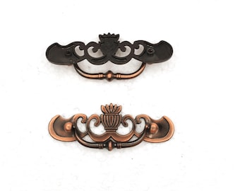 May include: Two decorative bronze-colored drawer pulls. The top pull is a dark bronze, while the bottom pull is a lighter copper color. Both pulls have an ornate design with a central decorative element and curved handles.