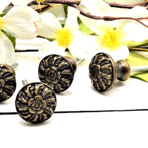 May include: Close-up of several antique bronze cabinet knobs with a floral design. The knobs have a dark bronze finish with gold highlights. White flowers and greenery are in the background.