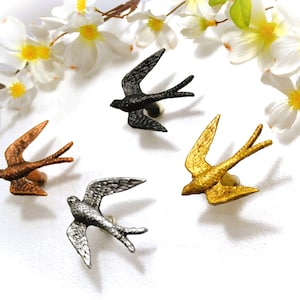 May include: A collection of decorative bird-shaped pins in various metallic finishes: bronze, black, silver, and gold. Each pin features a swallow design with outstretched wings, perfect for adding a touch of nature-inspired flair to clothing or accessories.