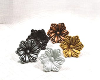 May include: A collection of five flower-shaped cabinet knobs in various finishes, including metallic silver, matte black, copper, gold, and a solid light gray. Each knob features a central circular button surrounded by six distinct, sculpted petals.