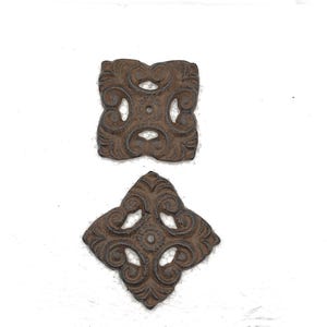 May include: Two decorative, cast iron pieces with an aged, brown finish. One is square, the other diamond-shaped, both featuring ornate, symmetrical scrollwork and openwork designs. These architectural elements are likely for furniture or home decor.