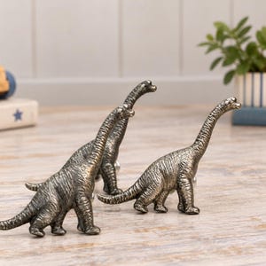 May include: Two metallic dinosaur-shaped drawer knobs with a dark, antique bronze finish. Each knob features a long-necked sauropod design with textured skin details, standing on four legs with a screw-in attachment point on the back.