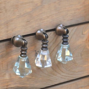 May include: Three antique-style cabinet knobs with clear, faceted glass crystal drops and bronze-toned metal hardware. The knobs are mounted on a wooden surface with visible wood grain.
