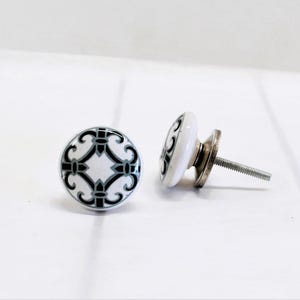 May include: Two white ceramic cabinet knobs with a black and gray decorative pattern. One knob is upright, and the other is lying on its side, showing the screw. The knobs are on a white surface.