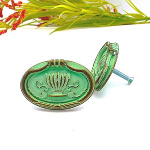 May include: Two oval-shaped, green cabinet knobs with a distressed finish and bronze-colored trim. Each knob features a decorative design and a silver screw for installation. The knobs are set against a white background.