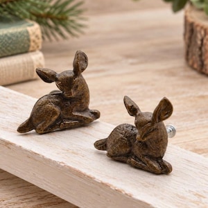 May include: Two bronze-colored cabinet knobs shaped like resting deer. Each knob features a detailed, textured finish depicting a fawn in a lying position with ears pointed upward. The knobs include a metal screw attachment on the back side.