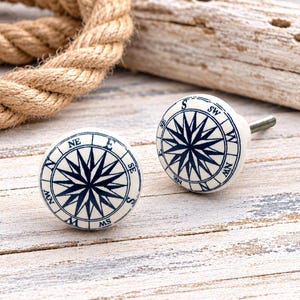 May include: Two round ceramic cabinet knobs featuring a dark blue compass rose design on a white background. Each knob has a metal screw post extending from the back, with cardinal directions N, S, E, and W printed around the edge.