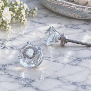 May include: Two clear glass cabinet knobs with a faceted, diamond-like shape. Each knob features a silver-toned metal bolt extending from the back, designed for attachment to furniture, complete with a small washer and nut for secure installation.