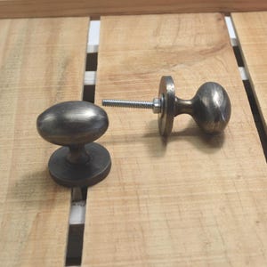 May include: Two antique-style bronze cabinet knobs with an oval shape. One knob is assembled with a threaded bolt, washer, and nut. The knobs are set against a light-colored wooden surface.