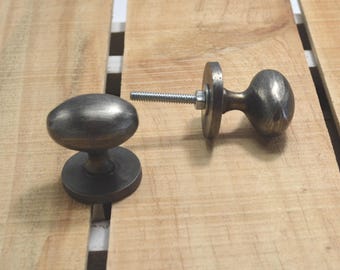 May include: Two antique-style bronze cabinet knobs with an oval shape. One knob is assembled with a threaded bolt, washer, and nut. The knobs are set against a light-colored wooden surface.