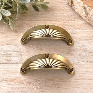 May include: Two semi-circular drawer pulls made of antique brass-toned metal. Each pull features a fan-shaped, ribbed design with three visible screw holes, one at the top center and two at the bottom corners, secured with matching screws.