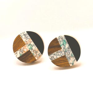 May include: A pair of round stud earrings. Each earring features a geometric design with sections of dark wood, black, and a speckled white and colored pattern. The earrings are set against a white background.