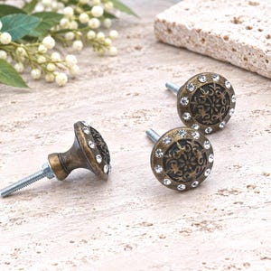 May include: Three round cabinet knobs with an antique brass finish and a dark center featuring an embossed floral pattern. Each knob is bordered by a ring of small, clear, sparkling crystals and includes a threaded metal bolt for installation.