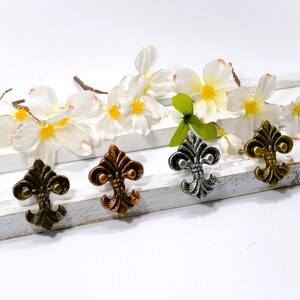 May include: Four decorative fleur-de-lis cabinet knobs in antique bronze, copper, silver, and gold finishes. The knobs are arranged on a white wooden surface, accented by white flowers with yellow centers.