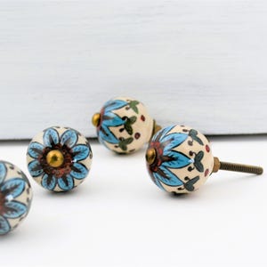 May include: A collection of ceramic cabinet knobs with a floral design. The knobs feature a cream-colored base with a blue and brown flower pattern and brass hardware. The knobs are decorative and suitable for furniture.