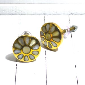 May include: Two decorative cabinet knobs. Each knob features a gold-colored circular base with a floral design made of white and cream-colored inlays. The knobs have a clear stem with a nut and bolt for installation.