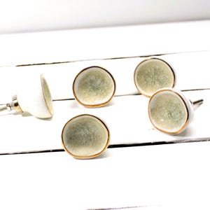 May include: Five decorative cabinet knobs. Each knob features a white base with a light green, textured face, and a gold-colored trim. The knobs are arranged on a white, wooden surface.