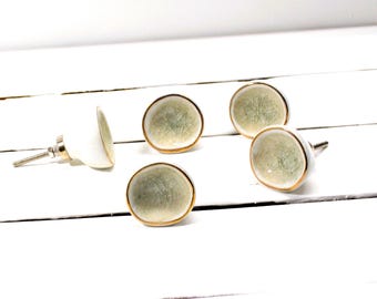 May include: Five decorative cabinet knobs. Each knob features a white base with a light green, textured face, and a gold-colored trim. The knobs are arranged on a white, wooden surface.