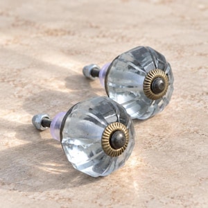 May include: Two clear, faceted glass cabinet knobs with a round, pumpkin-like shape. Each knob features a central antique brass-toned metal cap with a ridged edge, a threaded metal bolt, and a small clear plastic washer at the base.
