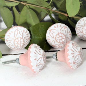 May include: A collection of decorative ceramic cabinet knobs. The knobs feature a white floral design on a pink base. The knobs are on a white wooden surface with green leaves in the background.