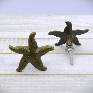 May include: Two bronze-colored starfish cabinet knobs. The knobs are in a five-point star shape with a textured surface. One knob is lying flat, and the other is upright with a screw and washer attached. The knobs are on a white wooden surface.