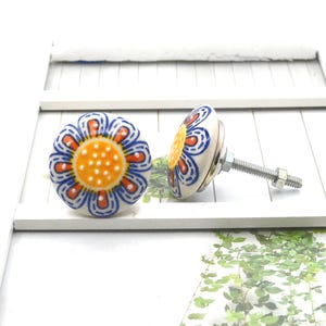 May include: Two ceramic cabinet knobs with a floral design. The knobs feature a white base with a blue, orange, and yellow flower pattern. Each knob has a metal screw for installation. The knobs are on a white surface.
