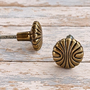 May include: Two round cabinet knobs with an antique brass finish. Each knob features a raised, radiating sunburst or shell-like pattern with deep grooves. One knob is shown from the side with its attached metal mounting screw.