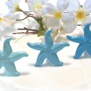 May include: Three light blue starfish figurines on a white plate, with white flowers and brown stems in the background. The starfish are a vibrant, solid color and have five points. The overall theme is coastal or nautical.
