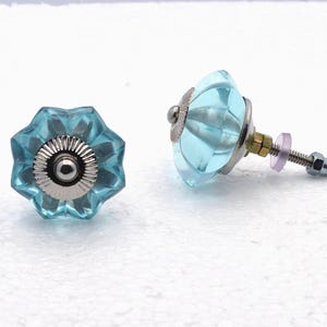 May include: Two light blue glass cabinet knobs with a starburst design. The knobs have a silver metal base and a screw with a nut for installation. The glass has a faceted, jewel-like appearance, adding a decorative touch to furniture.