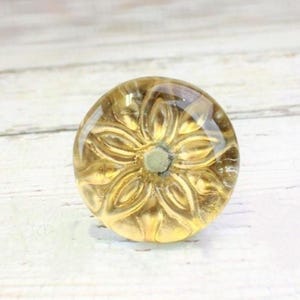 May include: A clear, golden-yellow glass cabinet knob with a floral design. The knob has a rounded shape and is mounted on a white wooden surface. The floral pattern is visible through the transparent glass.