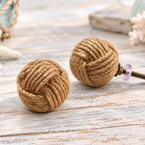 May include: Two spherical cabinet knobs crafted from natural tan jute rope wound into a tight knot. One knob features a metal bolt extending from the back with a small translucent purple washer and a silver-toned nut attached to the threaded end.