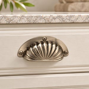 May include: A semi-circular metal cup pull handle with a scalloped, shell-like design and a brushed nickel finish. The handle is attached to a light-colored wooden drawer front with two visible screws on the left and right sides.