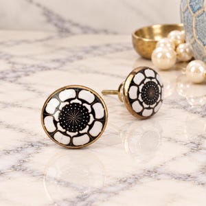 May include: Two round cabinet knobs featuring a black and white floral pattern with a central dotted design. Each knob has a smooth, glossy finish and a metallic gold-toned rim, with a short metal screw extending from the back for installation.