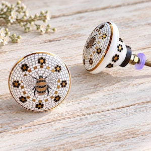 May include: Two round ceramic cabinet knobs featuring a white grid pattern, a central bee illustration, and small black and gold flower motifs. The knobs have a gold-toned rim and a metal screw attachment with a small purple plastic washer.