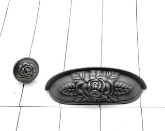 May include: A set of two antique-style cabinet hardware pieces. The set includes a round knob and a pull with a rose design. The hardware is made of a dark gray metal and is set against a white wood background.