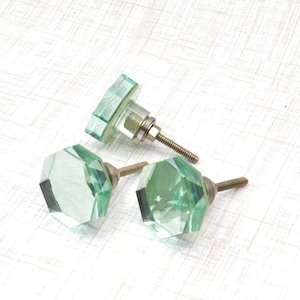 May include: Three light green, faceted glass cabinet knobs with brass hardware. The knobs have a geometric design and are arranged on a white surface. The knobs are a decorative element for furniture.