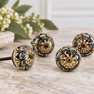 May include: Four round ceramic cabinet knobs featuring a cream-colored base with intricate black line work and metallic gold accents in a floral pattern. Each knob has a central bronze-toned metal screw and a rounded, smooth surface.