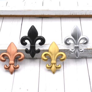 May include: Four fleur-de-lis decorative accents in copper, black, gold, and silver. The fleur-de-lis is a stylized lily, used as a decorative motif. The accents are displayed on a white wooden surface.