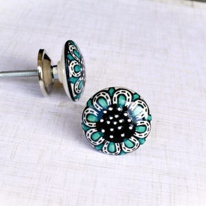 May include: Two decorative ceramic cabinet knobs. The knobs feature a floral design with teal and white accents on a black background. Each knob has a silver metal base and screw for easy installation.