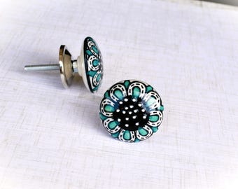 May include: Two decorative ceramic cabinet knobs. The knobs feature a floral design with teal and white accents on a black background. Each knob has a silver metal base and screw for easy installation.