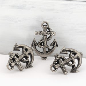 May include: Three silver-colored anchor-shaped decorative objects. The central anchor is upright, with two smaller anchors on either side. Each anchor features rope detailing and a classic design, suitable for nautical-themed decor.