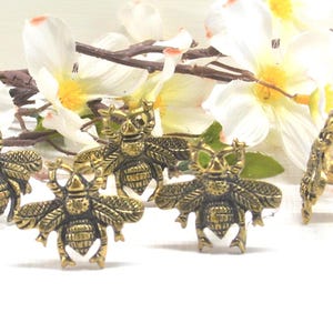 May include: Antique brass bee-shaped cabinet knobs. The knobs feature detailed wings and a textured body. The image shows several knobs with a floral arrangement in the background. The knobs are designed for furniture and home decor.