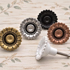 May include: A collection of five sunflower-shaped cabinet knobs in various finishes including antique gold, silver, black, copper, and white. Each knob features a central circular disc surrounded by detailed petals and a metal screw.