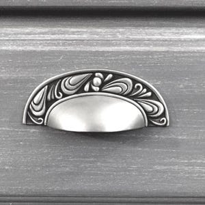 May include: A silver-toned, half-moon cabinet pull with an ornate, floral design. The pull is mounted on a gray, textured surface, and features a curved handle with detailed scrollwork and a central, rounded element.