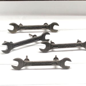 May include: Five metal wrench-shaped cabinet pulls. Each pull is a dark gray color and has the words "CHROME VANADIUM" engraved on it. The pulls are mounted on a white surface.