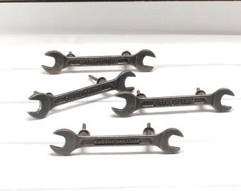 May include: Five metal wrench-shaped cabinet pulls. Each pull is a dark gray color and has the words "CHROME VANADIUM" engraved on it. The pulls are mounted on a white surface.