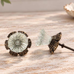 May include: Two cabinet knobs featuring clear, fluted glass tops shaped like flowers. Each knob has an ornate, antique bronze-colored metal base with intricate scrollwork details and a threaded metal bolt extending from the back.