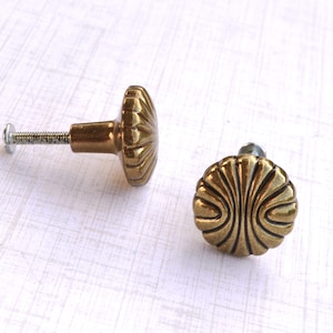 May include: Two antique brass cabinet knobs with a fan-shaped design. One knob is attached to a screw, while the other is separate. The knobs have a vintage aesthetic and are likely used for furniture or decorative purposes.