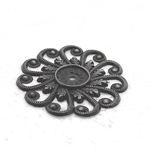 May include: A decorative, dark gray metal furniture rosette with an ornate, floral design. The rosette features a central circular element with a hole, surrounded by stylized leaves and swirling patterns. The overall shape is circular.